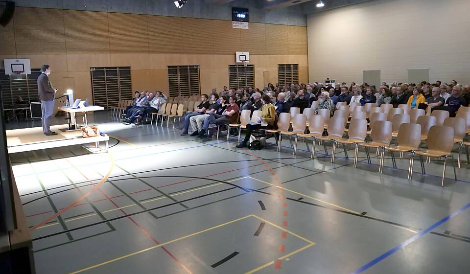 Rund 150 Personen sitzen in der abgedunkelten Mehrzweckhalle Kirchenfeld und hören den Ausführungen von Gemeindepräsident Ralph Lehner zu.